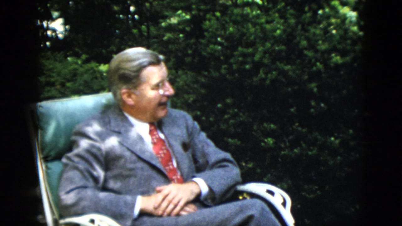Middle-aged man in grey flannel suit and red tie seated in a teal upholstered garden chair, hands clasped, dense foliage behind him.