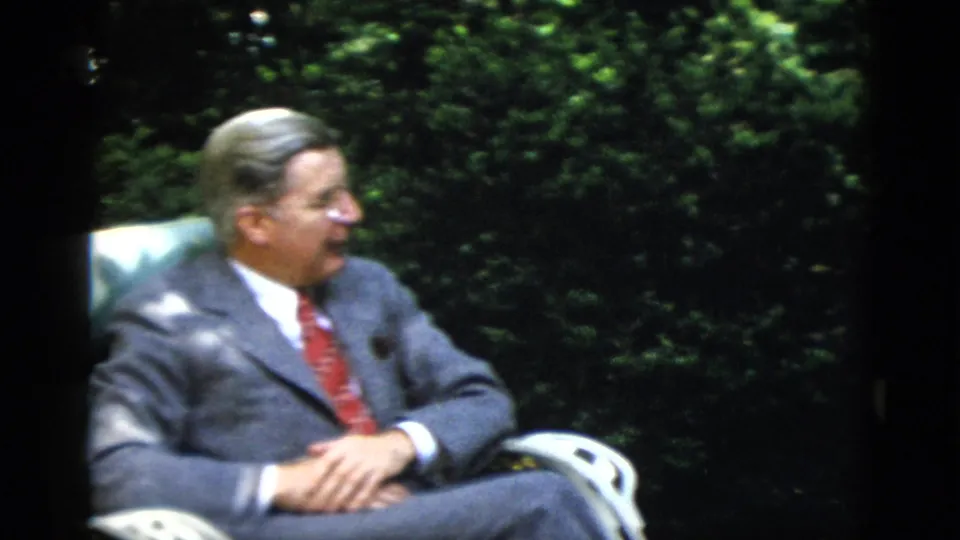 Unidentified man in 1950s grey flannel suit and red tie, relaxed and smiling in a teal garden chair, possibly at a Washington DC estate, circa 1952.