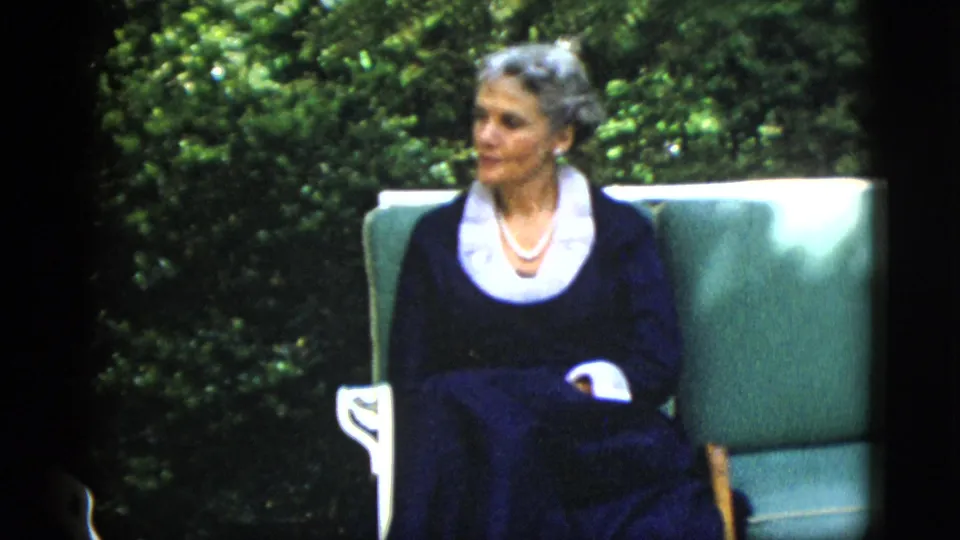 Unidentified woman in 1950s navy dress with white collar and pearl necklace, seated in a teal garden chair at what may be a Washington DC estate, circa 1952.
