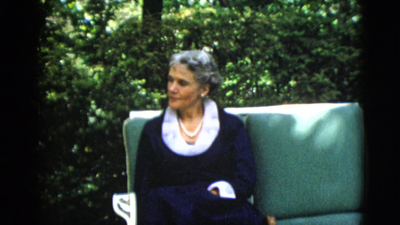 Older gray-haired woman in dark navy dress with white lace collar and pearl necklace, seated in a green cushioned outdoor chair with dense foliage behind.