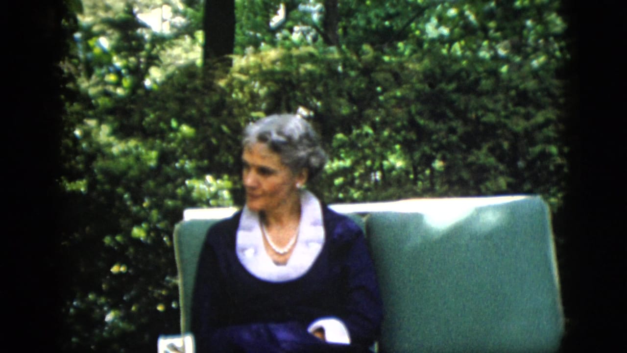 Woman with styled gray hair in dark navy dress with white lace collar and pearl necklace, glancing toward camera, seated in green cushioned garden chair; 1952 home movie.