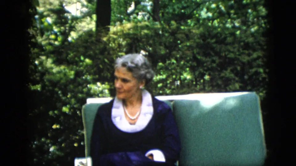 Woman with styled gray hair in dark navy dress with white lace collar and pearl necklace, glancing toward camera, seated in green cushioned garden chair; 1952 home movie.