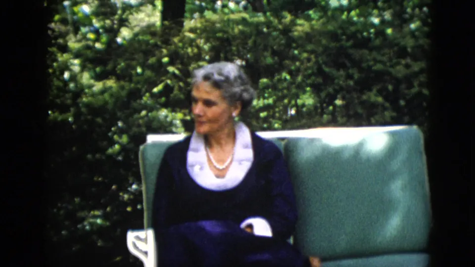 Older woman with gray hair in dark navy dress with white lace collar and pearl necklace, a slight smile, seated in green outdoor garden chair with dense foliage; 1952 home movie.