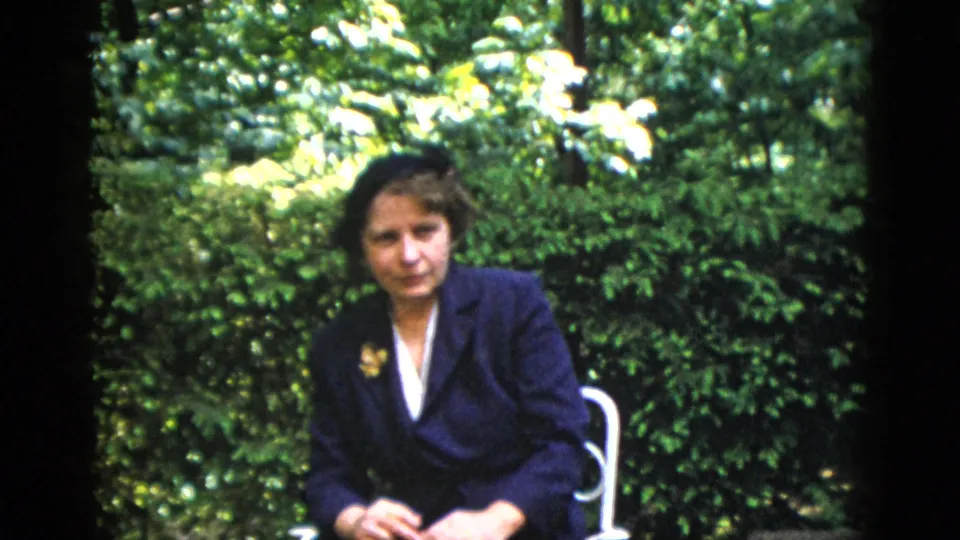 Dark-haired woman in structured navy blazer over white blouse with lapel brooch, seated in white metal garden chair with dense foliage behind; from a 1952 Washington DC home movie.