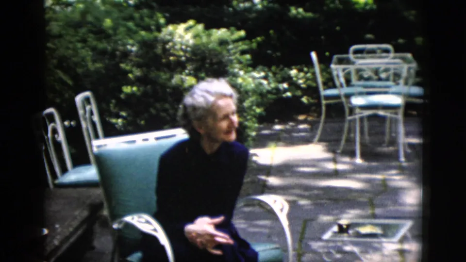 Elderly woman with white hair in dark clothing, in profile on garden patio, green metal chairs behind, circa 1952, 8mm home movie