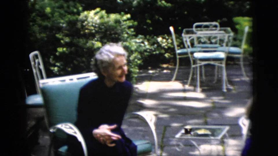 Elderly woman in dark clothing seated on garden patio, metal chairs and green garden backdrop, circa 1952 Washington DC, 8mm home movie