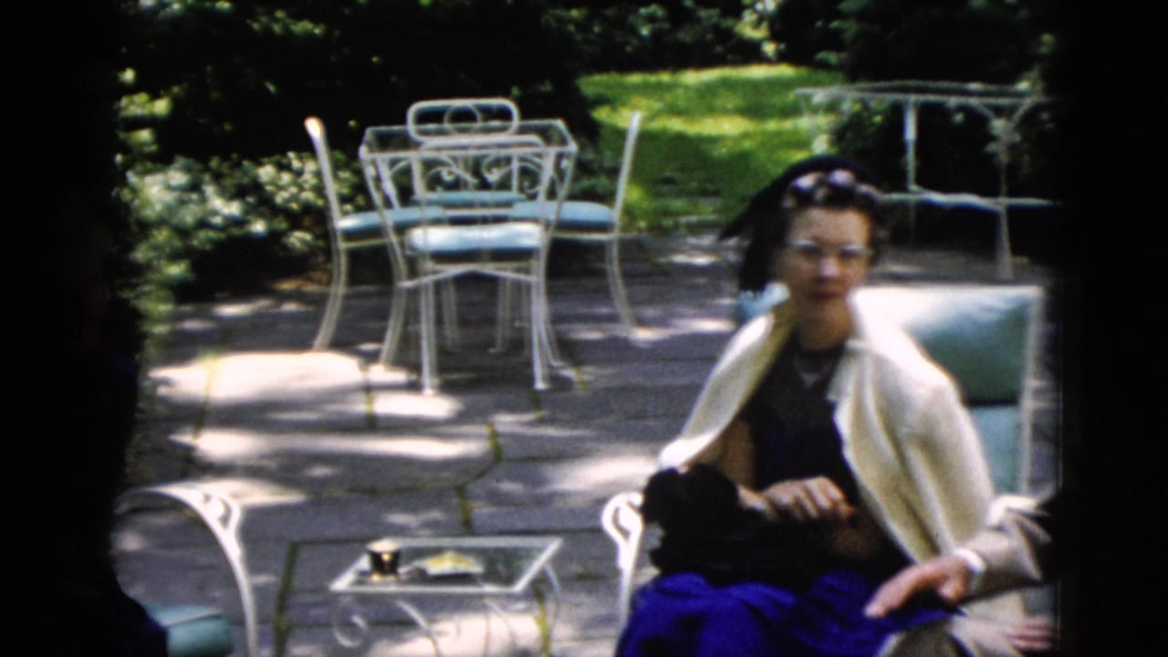 Woman in a dark dress and white shawl seated on garden patio furniture, a small dark dog visible in her lap.