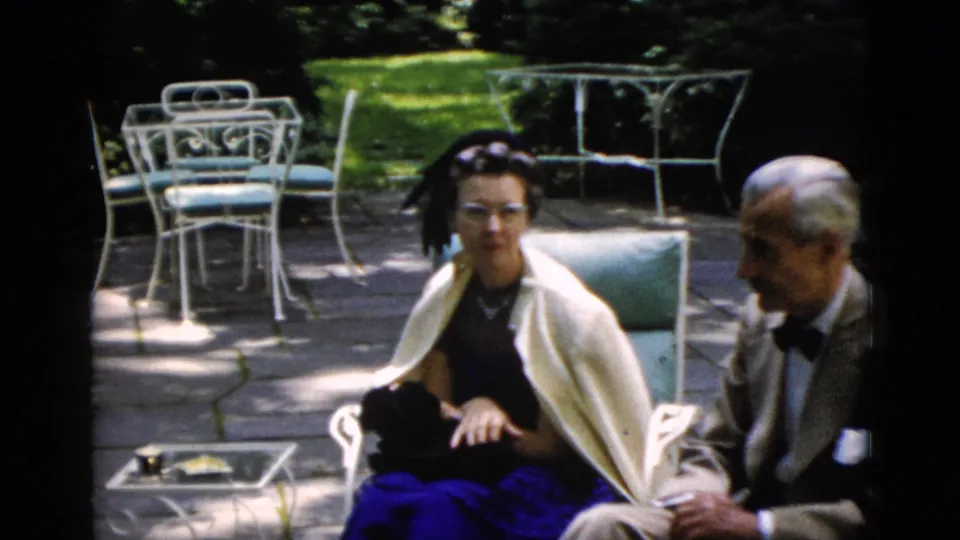 Older man in a tan suit with bow tie and younger woman in a dark dress conversing on a garden patio, Washington DC area, circa 1952.