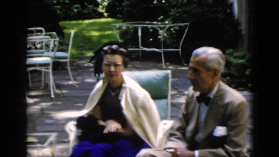Man in a tan suit and bow tie conversing with a woman in a dark dress and shawl on a garden patio, Washington DC area, circa 1952.