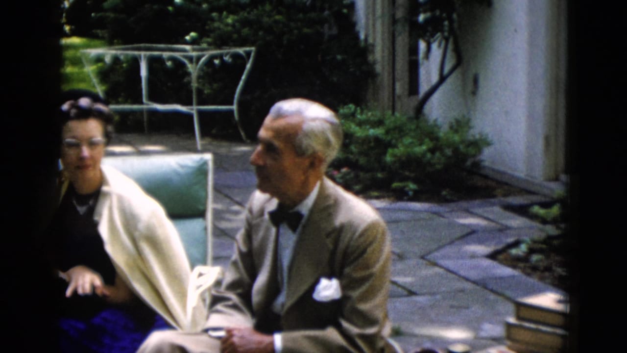 Older couple in tan suit and cream shawl continuing animated conversation on stone garden patio, wrought iron furniture behind them