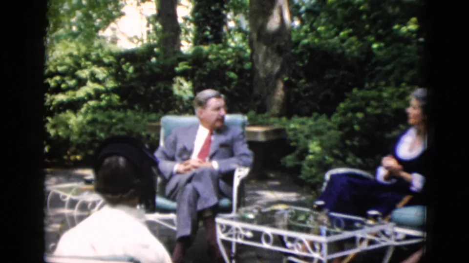 Group of adults in mid-century formal wear gathered around a white wrought iron table on a stone patio with a lush wooded garden backdrop, circa 1952