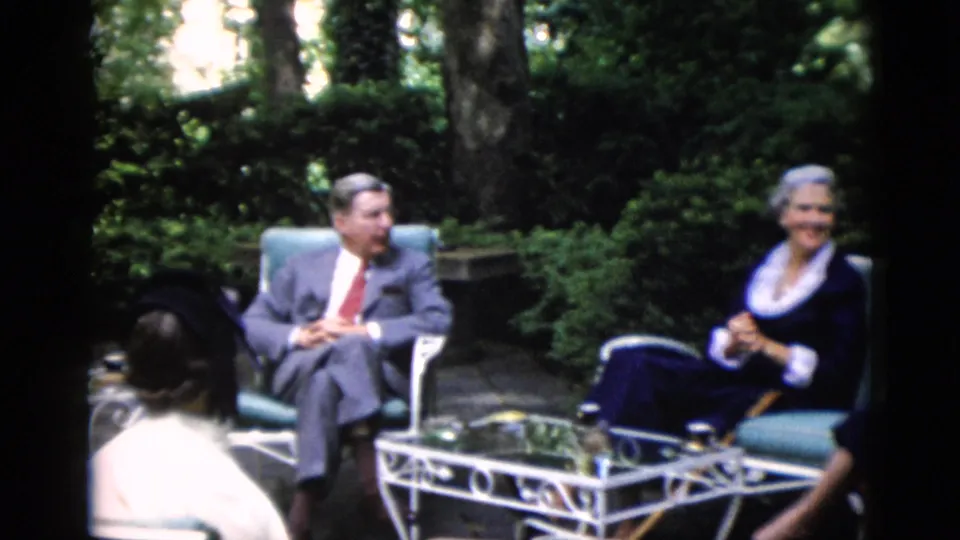 Man in grey suit with red tie converses with women in formal wear on a lush garden patio with white wrought iron furniture, circa 1952 Washington DC area