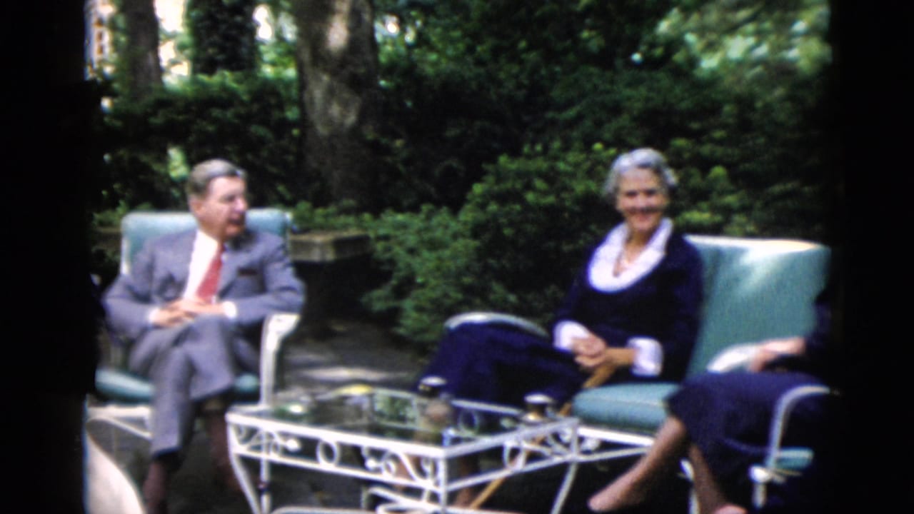 Man in gray suit and two women in navy dresses seated around a white wrought-iron patio table in a garden.