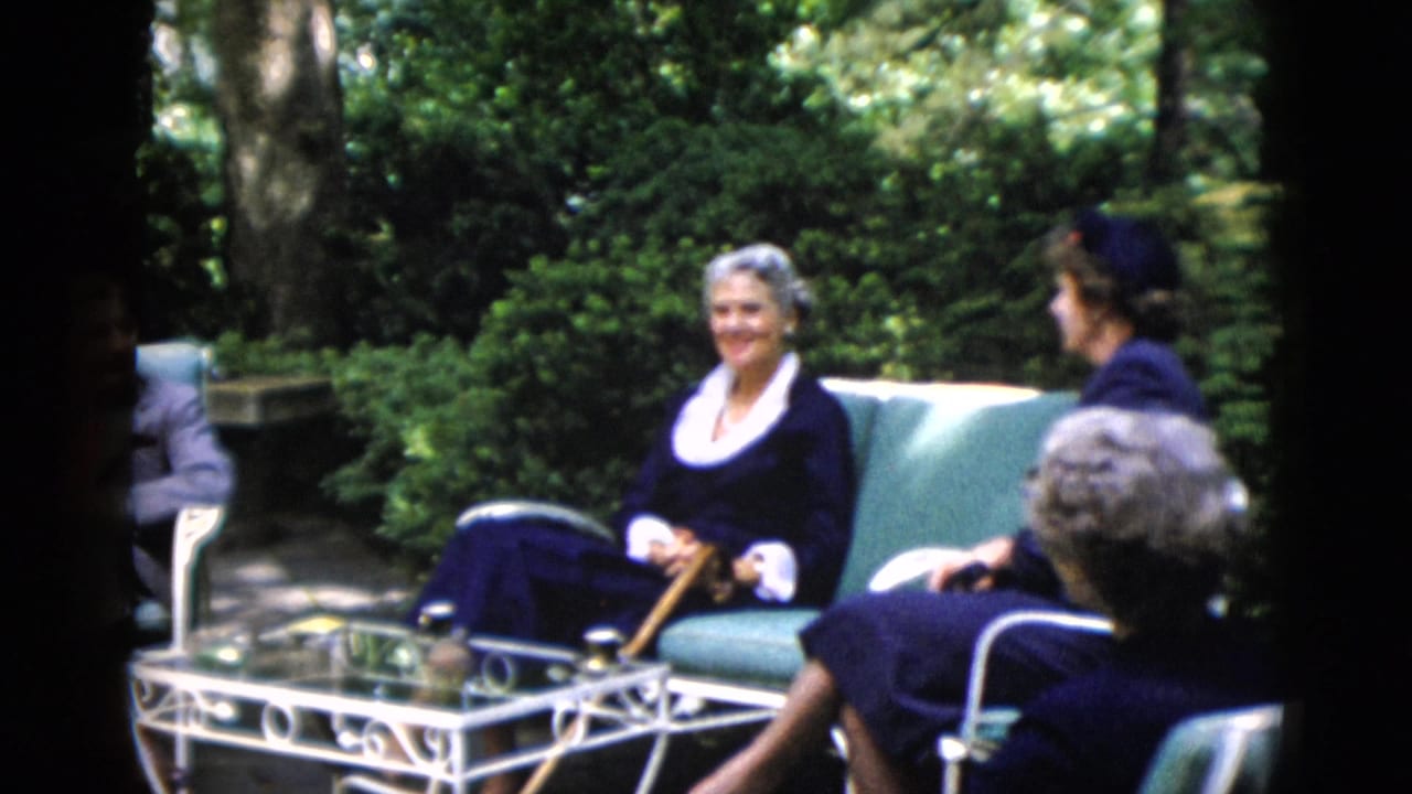 Older white-haired woman in navy dress and second woman in dark clothing seated in garden patio chairs; man partially visible at left edge.