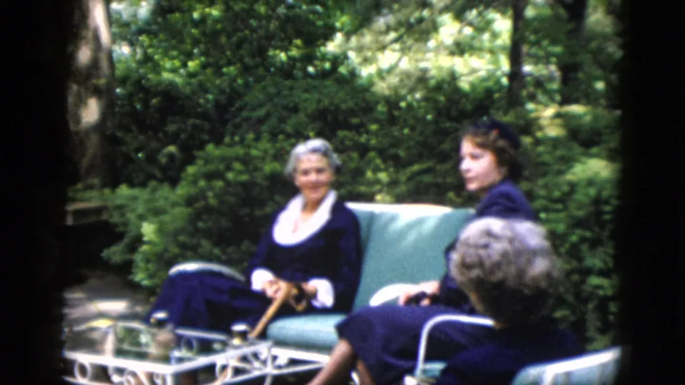 Three adults in outdoor garden gathering, 8mm home movie, circa 1952; foreground figure seen from behind holding a cane.