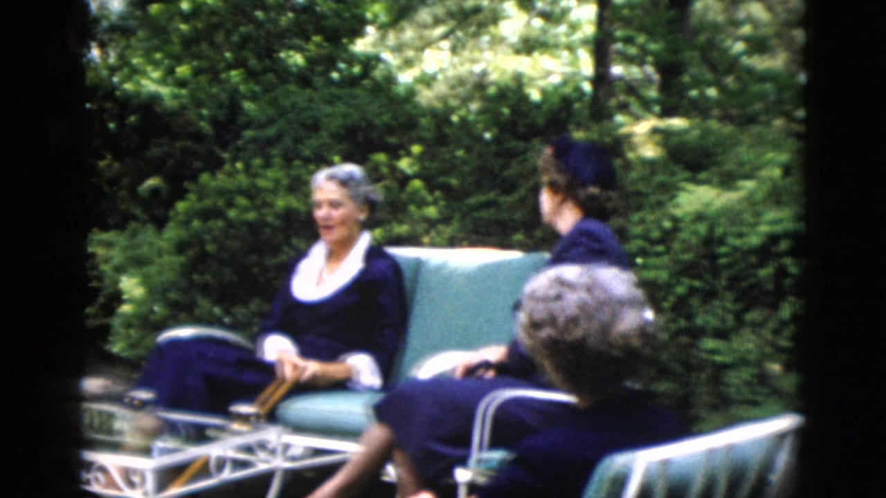 Two adult women in early-1950s dark navy dresses on green metal garden chairs amid dense summer foliage; from a home movie reportedly shot in Washington DC in 1952.