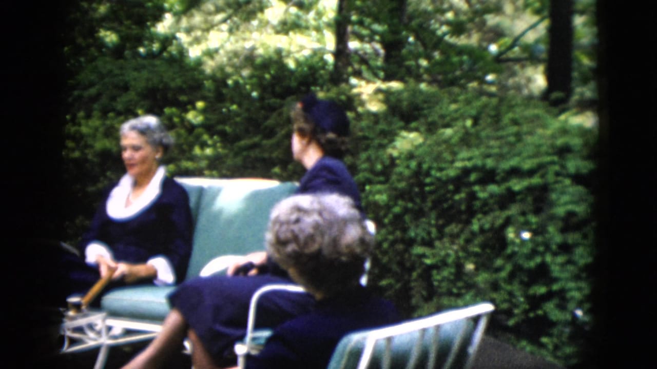 Same two women on garden furniture; the rear-facing woman wears a dark hat, the forward-facing woman has white hair and a white-collared dark dress.