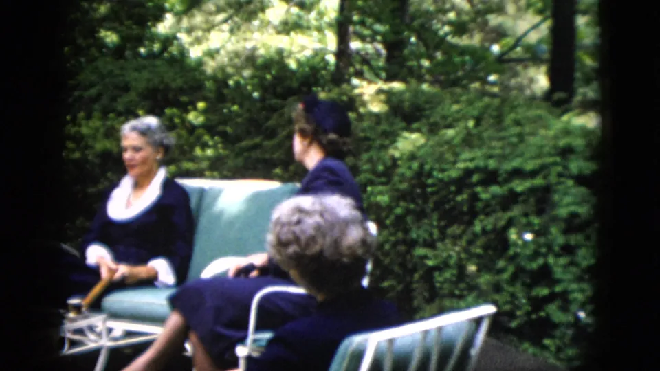 Two adult women in early-1950s attire on green metal garden chairs amid lush summer trees; from a 1952 home movie reportedly shot in Washington DC.