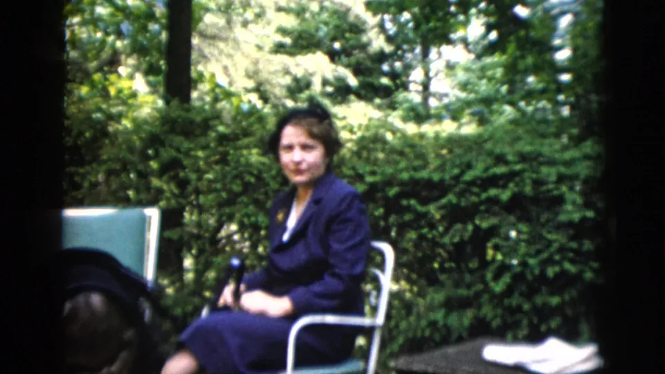 Young adult woman in dark clothing seated in a white metal garden chair surrounded by dense summer foliage; from a 1952 home movie reportedly shot in Washington DC.