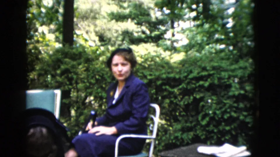 Close view of a dark-haired adult woman in dark clothing on metal garden furniture with dense leafy trees behind her; from a 1952 home movie reportedly shot in Washington DC.