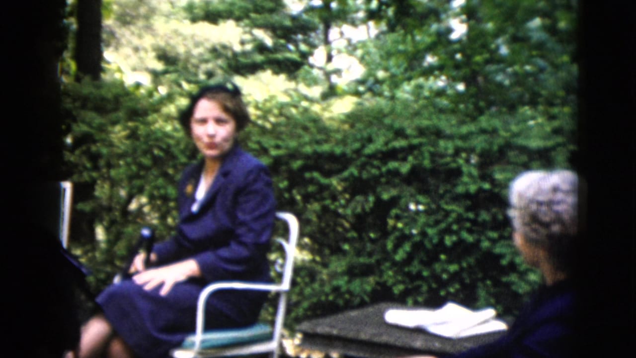Woman in dark navy suit seated in white tubular-metal garden chair, green hedge backdrop, dark patio table with papers visible at right.