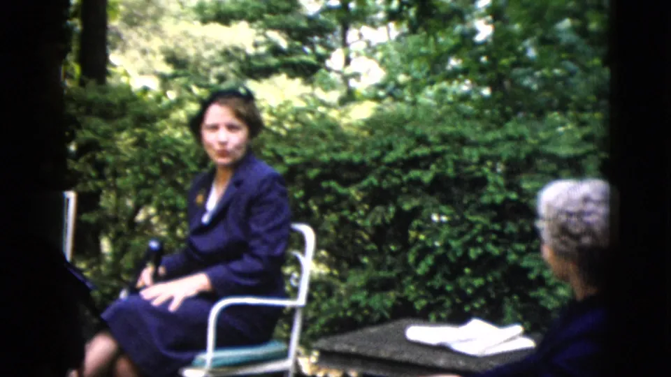 Dark-haired woman in dark navy suit in white tubular-metal garden chair on a patio, c. 1952, likely Washington DC; lush deciduous hedge behind.