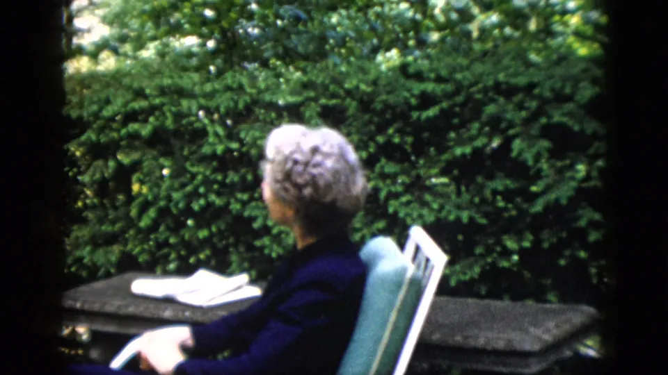 Older woman with gray hair seen from behind in teal metal garden chair on a private garden patio, papers on table beside her, c. 1952.