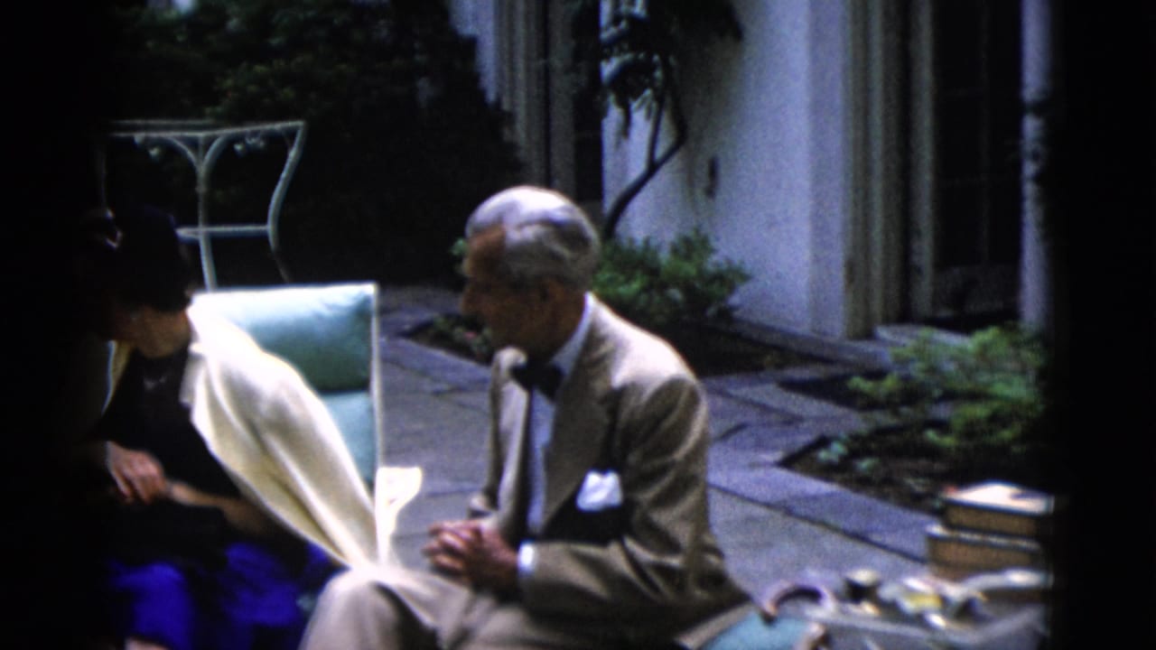 Older man in tan suit with dark bow tie and a woman in bright blue skirt and cream shawl sit in light-blue cushioned chairs on a flagstone patio, circa 1952, Washington DC.