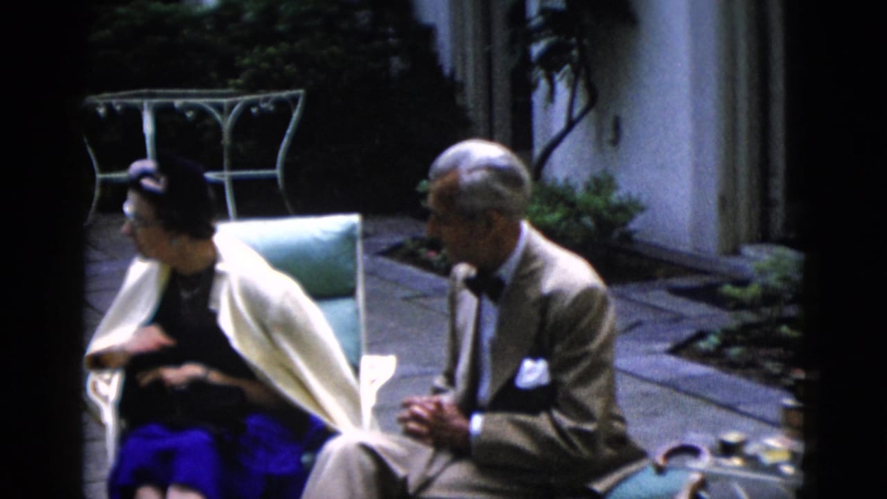 Man in tan suit with bow tie and woman wearing a dark hat and blue skirt seated in garden chairs on a stone patio surrounded by greenery.