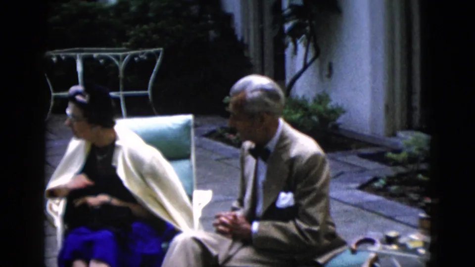 Man in tan suit and dark bow tie with a woman in a dark hat and bright blue skirt sit in light-blue garden chairs on a flagstone patio, circa 1952, Washington DC.