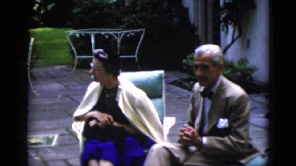 Older woman wearing glasses, bright blue skirt, and cream shawl seated beside an older man in tan lounge suit and dark bow tie on a flagstone patio, circa 1952.