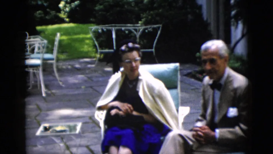 Wide patio view showing an older couple in mid-century dress seated in light-blue garden chairs, with white wrought-iron furniture and a manicured lawn beyond, circa 1952, Washington DC.