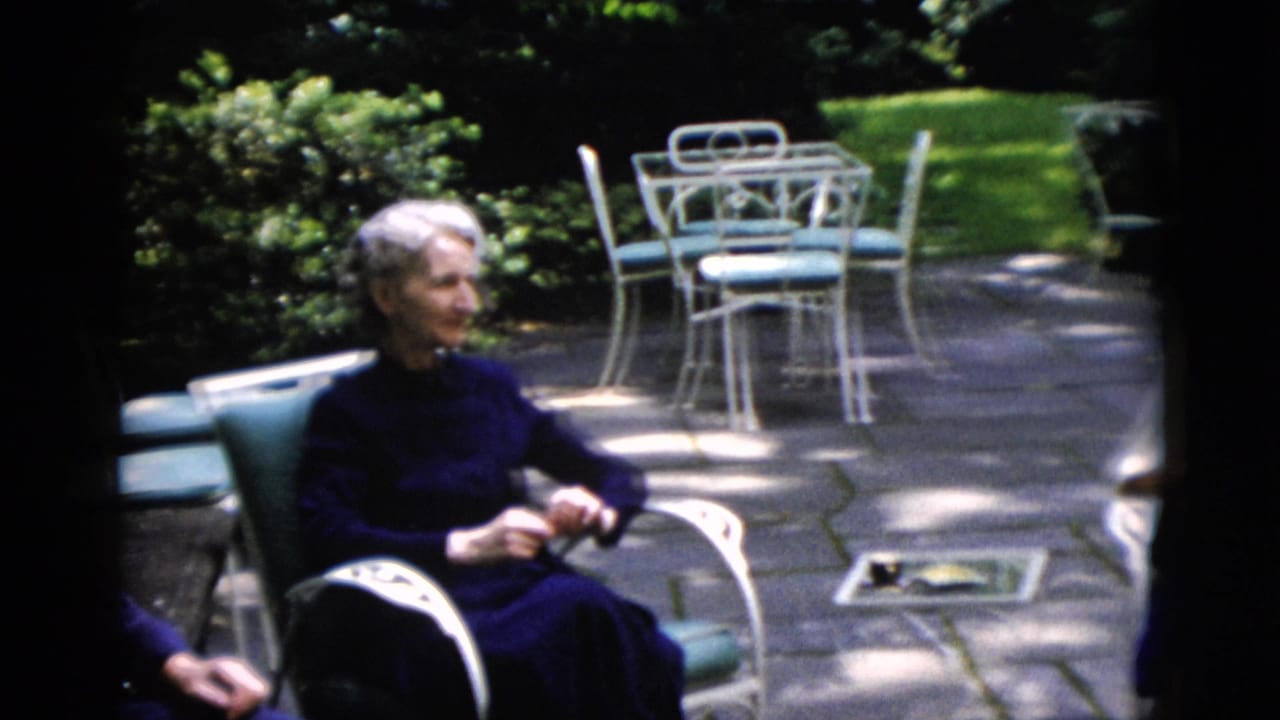 Older woman in a dark dress sits on a patio near white metal furniture in Washington, DC home-movie footage from 1952.