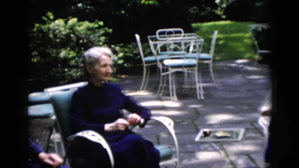Older woman in a dark dress sits on a patio near white metal furniture in Washington, DC home-movie footage from 1952.