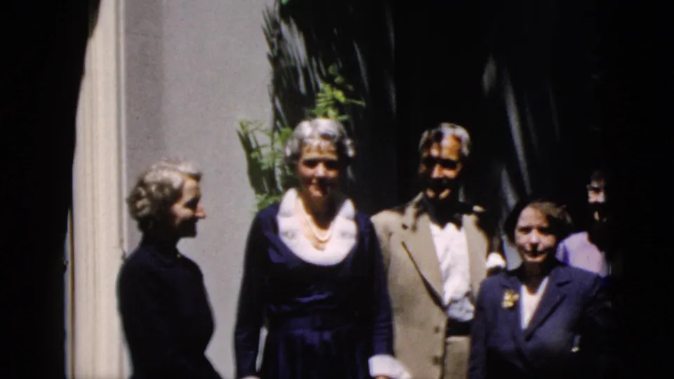 Adults pose outdoors beside a building, with a woman in a dark dress and a man in a light suit centered in the 1952 home movie.