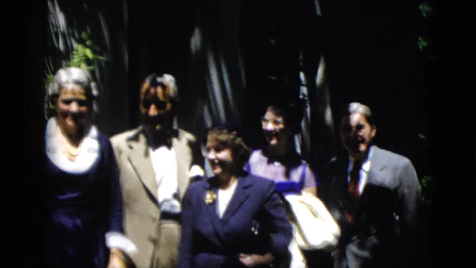 A 1952 Washington DC home-movie frame shows adults in formal period clothing standing together outdoors near greenery.