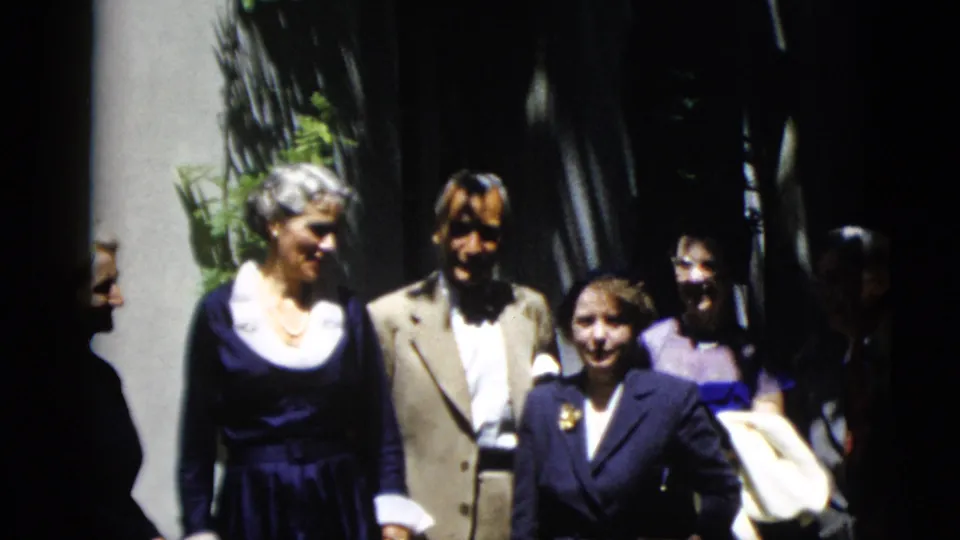 1952 Washington DC frame showing adults in formal clothing near a building edge and shaded foliage.