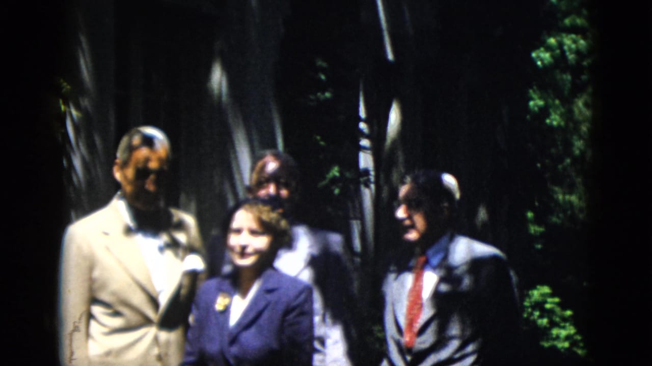 Formally dressed adults stand outdoors among tall trees in dappled sunlight.