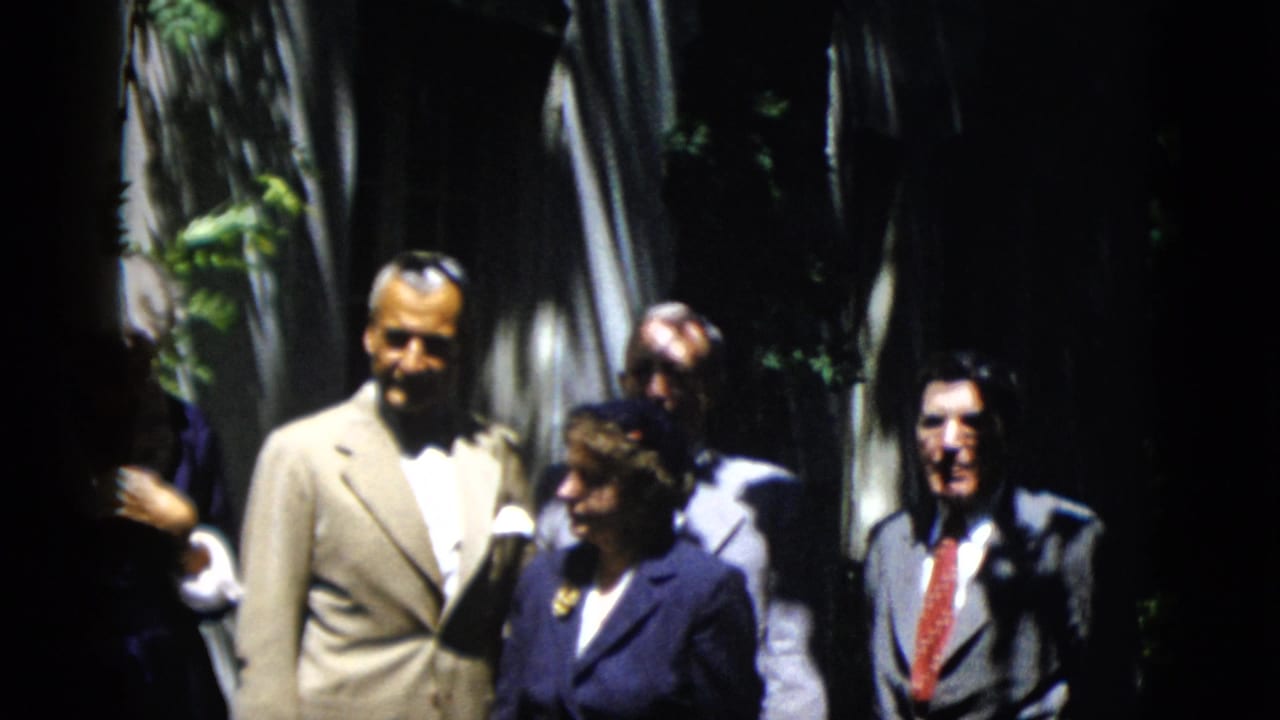 A man in a light suit and several adults stand close together under trees.