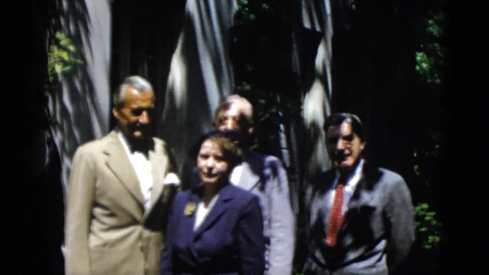 In 1952 Washington DC home-movie footage, adults in suits and dresses stand together in a shaded garden setting.