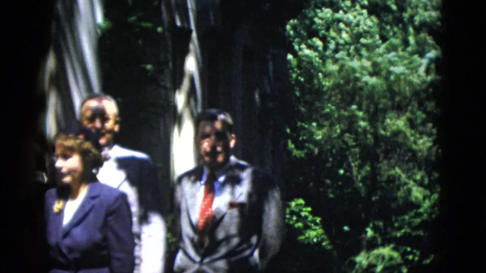 A woman and two men in formal dress stand in a shaded garden scene from 1952 Washington DC home-movie footage.