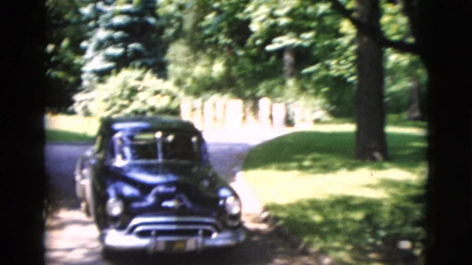 1952 Washington, DC home-movie frame showing a dark rounded sedan on a driveway amid trees and lawn.