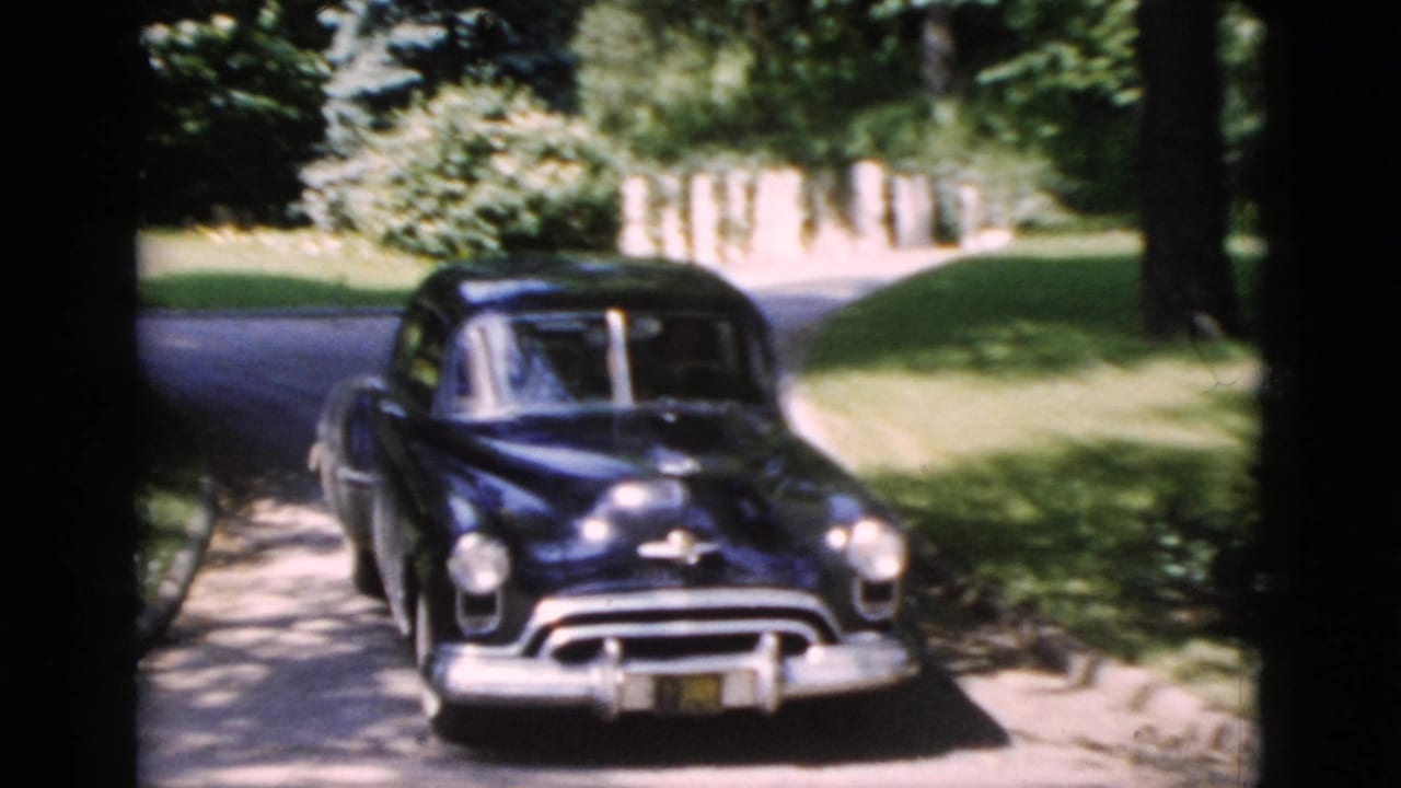 1952 Washington, DC home-movie frame showing a dark mid-century sedan on a shaded residential street with lawns and trees.
