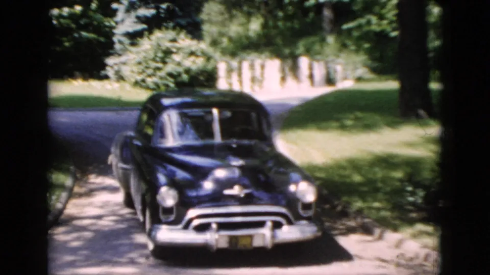 1952 Washington, DC home-movie frame showing a dark mid-century sedan on a shaded residential street with lawns and trees.
