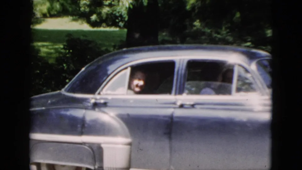 1952 Washington, DC frame showing the sedan in profile as a passenger looks out from the side window in a shaded residential setting.