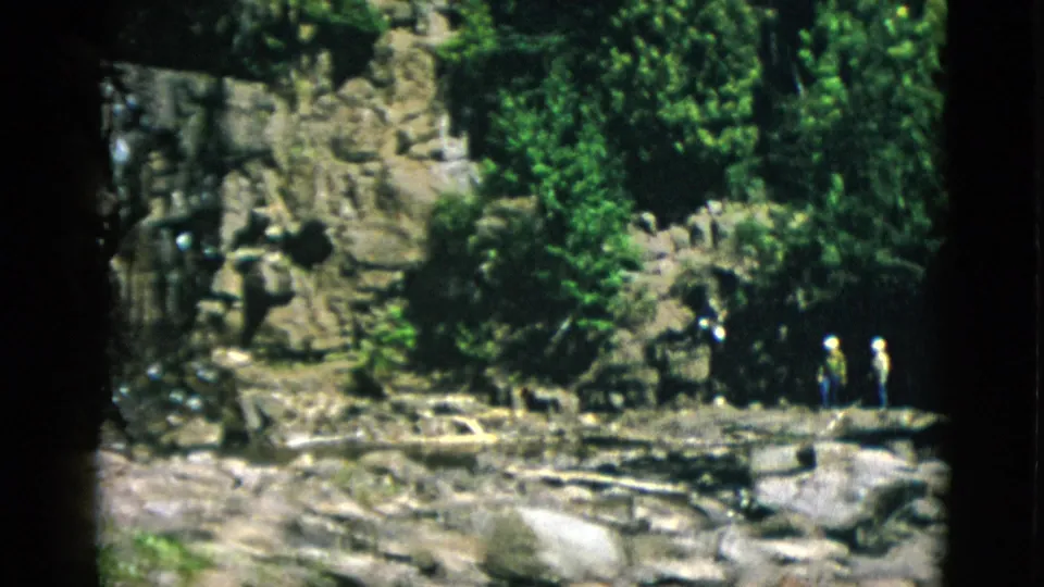 Two or more distant visitors appear near a rocky watercourse in the forested Quebec vacation scene.