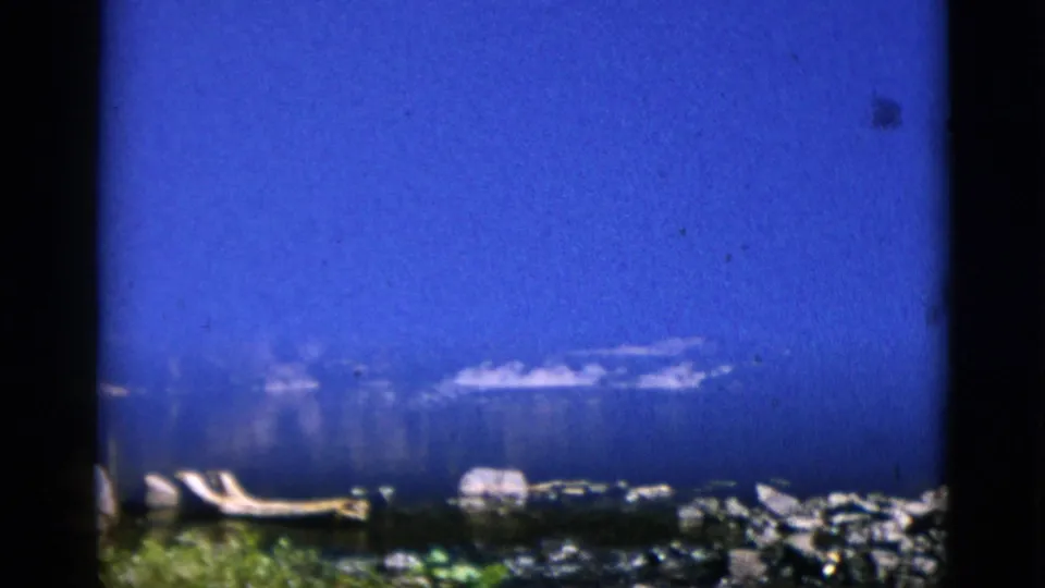 8mm home-movie frame from Quebec, Canada, circa 1952 — rocky shore under a heavily saturated blue sky, flat stones, and a light-coloured object near the waterline.