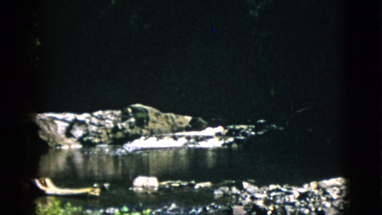 Dark contrasty frame showing a large rock outcrop above calm reflecting water, scattered stones in the foreground.