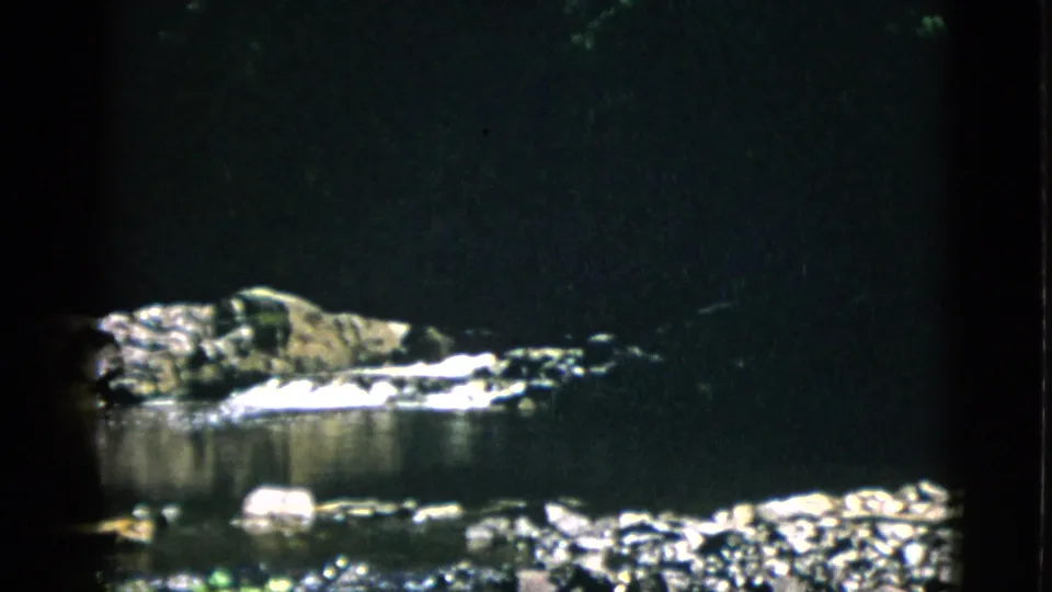 8mm home-movie frame circa 1952, Quebec — low rock mass above quiet water with heavy vignetting and strong contrast between lit rocks and surrounding shadow.