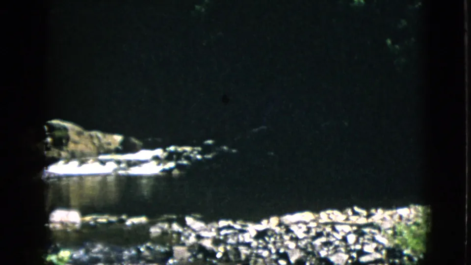 8mm home-movie frame circa 1952, Quebec — rock formation above still water, sunlit stones in foreground, heavy shadow framing typical of 8mm exposure limitations.